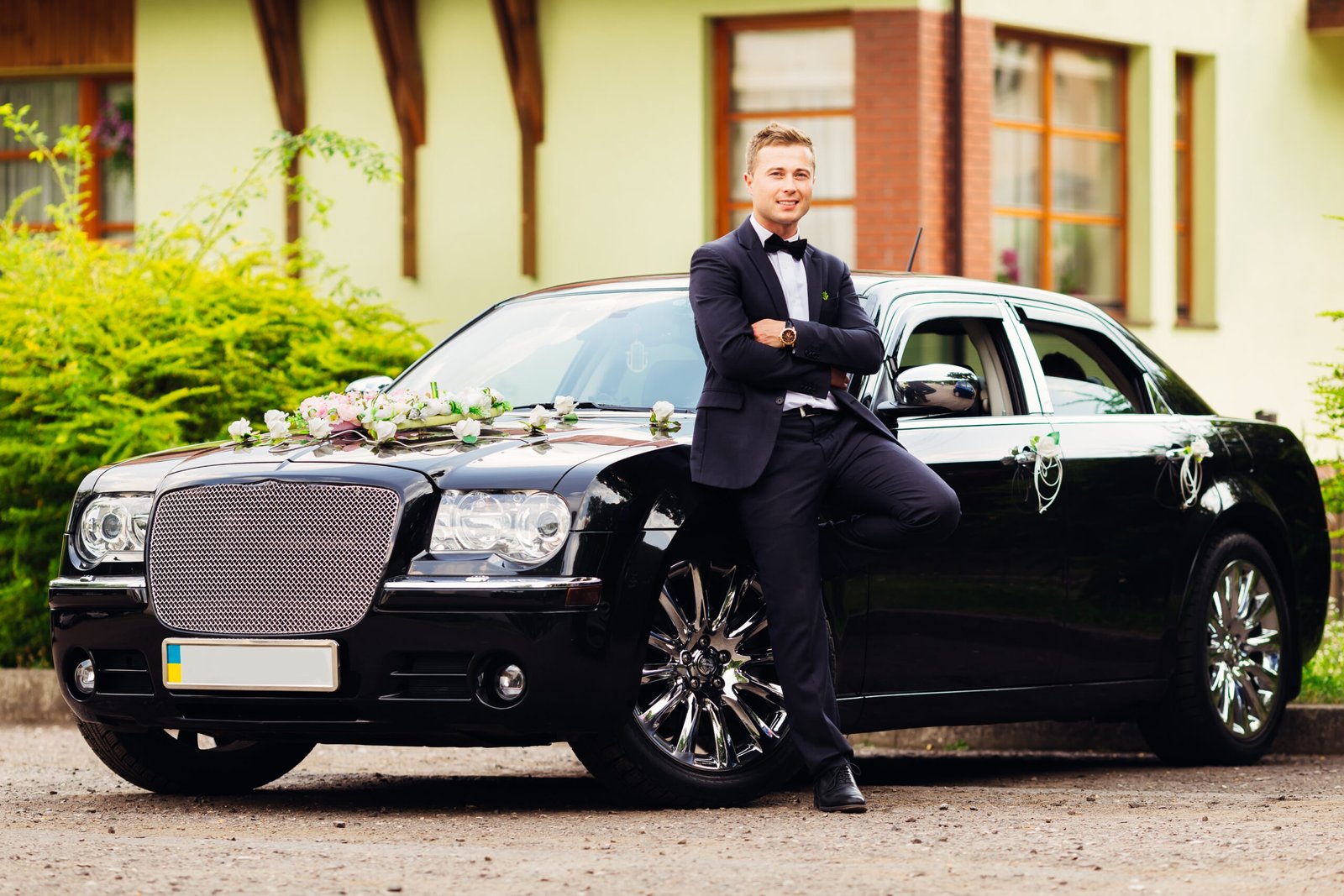 the fiance in a black suit stands near a beautiful black car